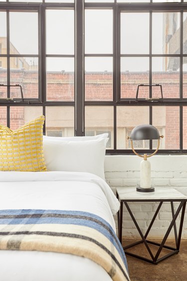 Modern bedroom with bed, white bedding, striped blanket, metal nightstand, metal and marble lamp, large industrial windows.